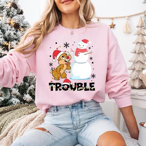 Puede incluir: Sudadera rosa de Navidad con un chipmunk de dibujos animados que lleva un gorro de Pap&aacute; Noel y sostiene una cadena de luces. El chipmunk est&aacute; de pie junto a un mu&ntilde;eco de nieve que lleva un gorro de Pap&aacute; Noel y una bufanda roja. El texto "Trouble" est&aacute; impreso debajo de la imagen.