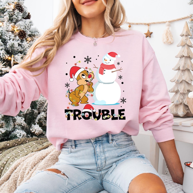 Puede incluir: Sudadera rosa de Navidad con un chipmunk de dibujos animados que lleva un gorro de Pap&aacute; Noel y sostiene una cadena de luces. El chipmunk est&aacute; de pie junto a un mu&ntilde;eco de nieve que lleva un gorro de Pap&aacute; Noel y una bufanda roja. El texto "Trouble" est&aacute; impreso debajo de la imagen.