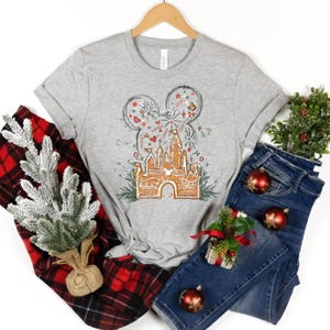 K&ouml;nnte beinhalten: Ein hellgraues T-Shirt mit einem festlichen Design, das Mickey Mouse-Ohren mit Weihnachtsschmuck und Feuerwerk zeigt, sowie ein Lebkuchenschloss. Das T-Shirt wird mit blauen Jeans, rotem Karomuster und Weihnachtsdekorationen kombiniert.