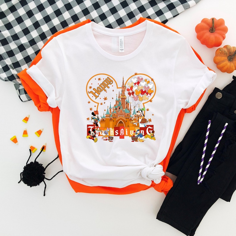 May include: White t-shirt with an orange trim featuring a Thanksgiving-themed graphic. The design includes a castle, Mickey Mouse, Minnie Mouse, and other characters, with the words "Happy Thanksgiving" in a festive font. The shirt is paired with black pants and decorative pumpkins.