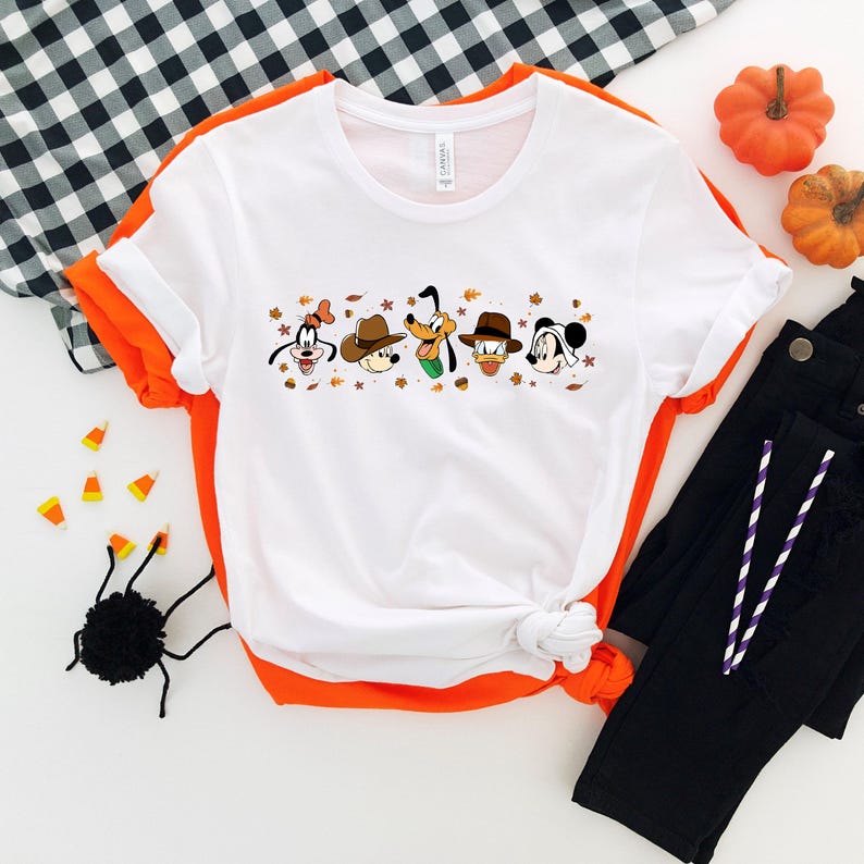May include: White t-shirt featuring cartoon characters in hats and autumn leaves. The shirt is styled with black pants, pumpkins, and Halloween decorations. The shirt has a round neck and short sleeves.