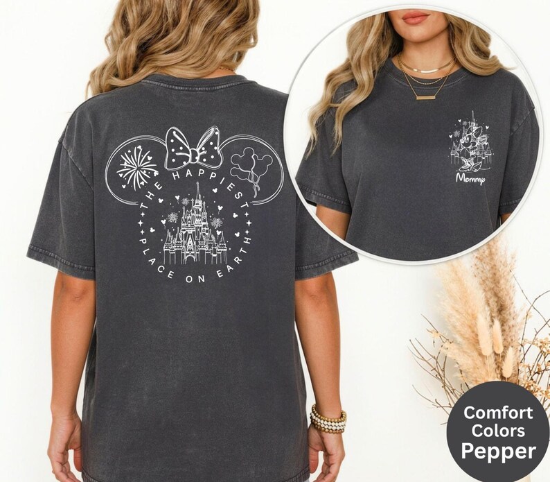 May include: Dark grey t-shirt with a white graphic on the back featuring a castle, fireworks, and the text "The Happiest Place on Earth." A smaller graphic on the front says "Mommy." The shirt is part of the Comfort Colors Pepper collection.