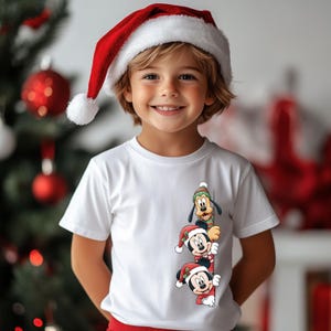 May include: A child wearing a white t-shirt with a Christmas-themed graphic featuring Mickey Mouse, Minnie Mouse, and Goofy. The child is also wearing a red Santa hat with a white trim and pom-pom. The background includes a Christmas tree.