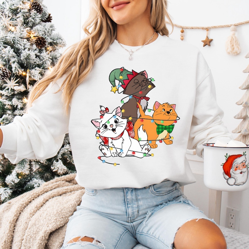 May include: White sweatshirt featuring a graphic of three cartoon cats adorned with Christmas lights and festive accessories. The cats are a mix of white, brown, and orange, with a Christmas tree in the background. A Santa mug is visible.