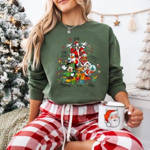 K&ouml;nnte beinhalten: Gr&uuml;nes Sweatshirt mit einem Weihnachtsbaum-Design mit Disney-Figuren wie Mickey Mouse und Donald Duck. Das Sweatshirt hat die Unterschriften der Charaktere und wird mit rot-wei&szlig; karierten Pyjamahosen getragen. Eine Tasse mit Santa-Design wird gehalten.
