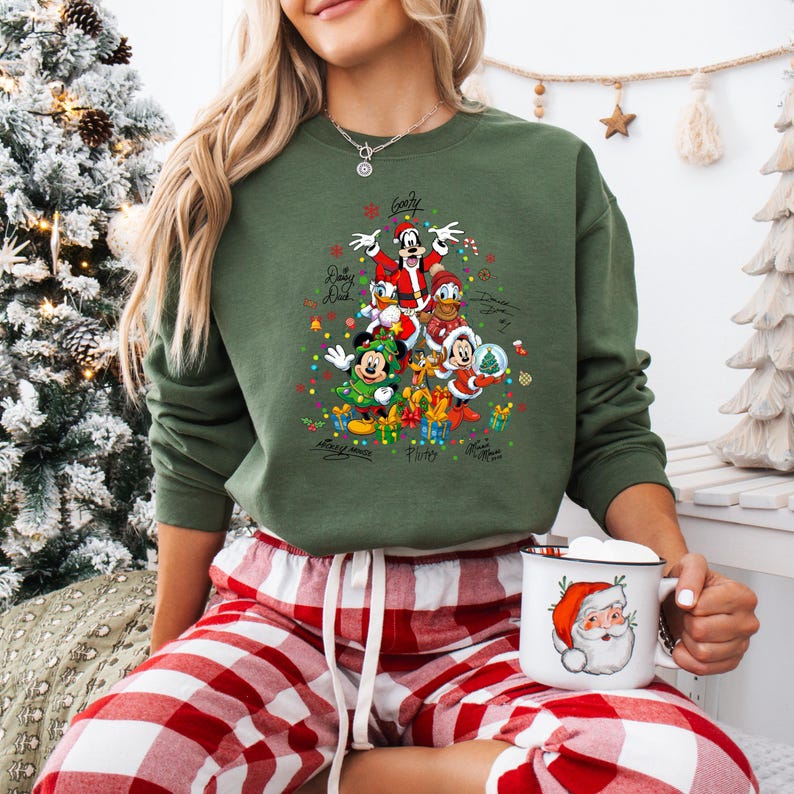 K&ouml;nnte beinhalten: Gr&uuml;nes Sweatshirt mit einem Weihnachtsbaum-Design mit Disney-Figuren wie Mickey Mouse und Donald Duck. Das Sweatshirt hat die Unterschriften der Charaktere und wird mit rot-wei&szlig; karierten Pyjamahosen getragen. Eine Tasse mit Santa-Design wird gehalten.