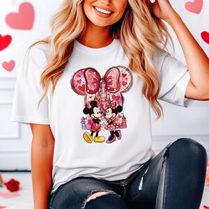May include: White t-shirt featuring a graphic of Mickey and Minnie Mouse holding hands in front of a castle. The design includes a large pink bow with heart accents and a pink and purple castle. The characters are in a loving embrace.