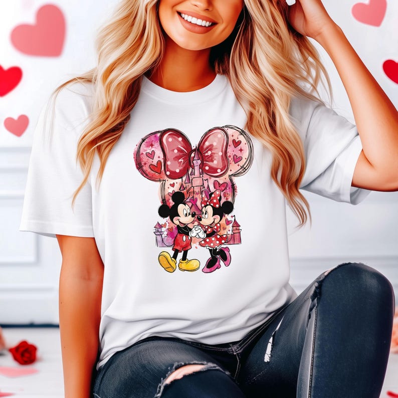 May include: White t-shirt featuring a graphic of Mickey and Minnie Mouse holding hands in front of a castle. The design includes a large pink bow with heart accents and a pink and purple castle. The characters are in a loving embrace.