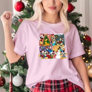May include: A light pink t-shirt featuring a colorful graphic of Disney characters celebrating Christmas. The graphic is divided into four sections, each showcasing different characters like Mickey Mouse, Minnie Mouse, and Donald Duck in festive scenes.