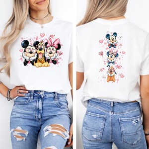 May include: White t-shirt featuring a colorful graphic of Mickey Mouse, Minnie Mouse, and Pluto. The front design shows the characters together, while the back has a vertical arrangement of the characters with hearts and stars.