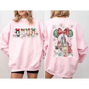 May include: A light pink sweatshirt featuring a watercolor design of Disney princesses. The front shows the princesses with Mickey Mouse ears. The back displays a castle with Mickey Mouse ears and a red bow.