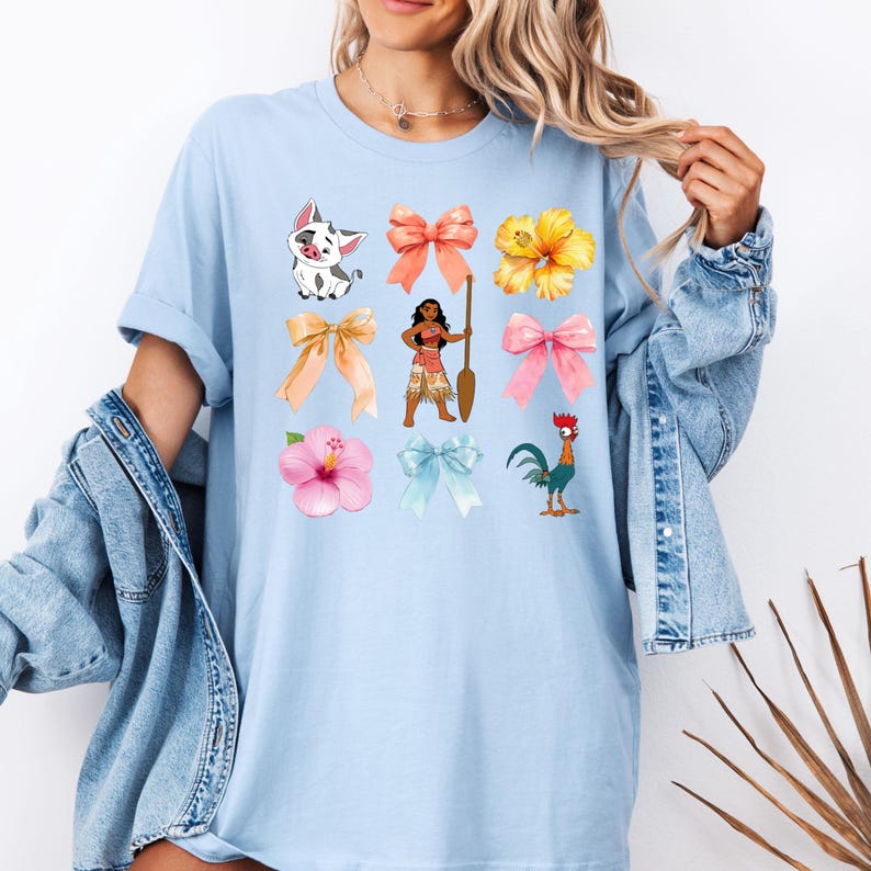 Pu&ograve; includere: Maglietta blu chiaro con una variet&agrave; di immagini di cartoni animati colorati, tra cui un maiale, un fiore, un gallo, un fiocco e una ragazza che tiene una pagaia.