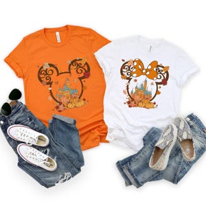 May include: Two t-shirts with a fall-themed Mickey Mouse and Minnie Mouse design. The Mickey Mouse shirt is orange and the Minnie Mouse shirt is white. Both shirts feature a silhouette of Mickey or Minnie's head with a castle inside and fall leaves around it.