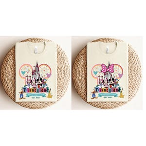 May include: Two cream colored t-shirts with a graphic print of Mickey Mouse and friends in front of the Disneyland castle. The text "Disneyland" is printed below the image. The text "The happiest place on earth" is printed below the Disneyland text. The text "Est. 1955" is printed below the previous text.