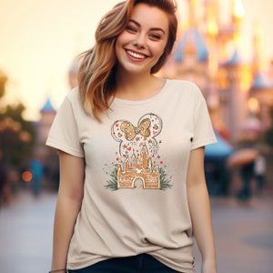 May include: A beige t-shirt with a graphic of a cartoon castle with a bow on top. The castle is surrounded by fireworks and greenery. The graphic is in a sketched style.