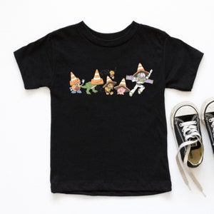 Op de afbeelding: Zwart T-shirt met een afbeelding van Toy Story-personages die verkeerskegels dragen. De personages zijn Woody, Buzz Lightyear, Rex, Mr. Potato Head en Hamm.