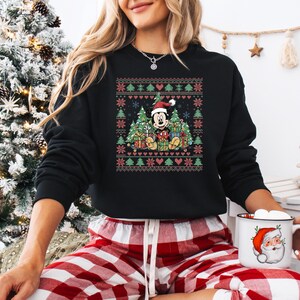 May include: Black sweatshirt featuring a festive Christmas design with Mickey Mouse wearing a Santa hat, surrounded by Christmas trees and gifts. The design incorporates red, green, and white elements, creating a holiday theme. The sweatshirt is paired with red and white plaid pajama pants.