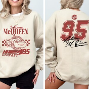May include: A beige sweatshirt with a red graphic of Lightning McQueen from the Cars movie. The graphic includes the text "Lightning McQueen", "Think Fast", and "95". The back of the sweatshirt has a red number "95" with the text "Lightning McQueen" in black.