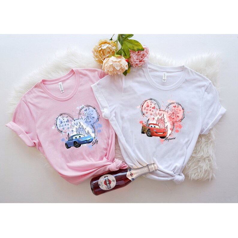 Puede incluir: Dos camisetas rosas y blancas con un dise&ntilde;o de cabeza de Mickey Mouse que presenta un coche de la pel&iacute;cula Cars. La camiseta rosa tiene un coche azul con la palabra "Sally" y la camiseta blanca tiene un coche rojo con la palabra " McQueen".