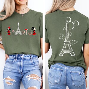 K&ouml;nnte beinhalten: Olivgr&uuml;nes T-Shirt mit wei&szlig;er Grafik des Eiffelturms und dem Schriftzug "PARIS". Mickey und Minnie sind auf der Vorderseite abgebildet. Die R&uuml;ckseite des T-Shirts zeigt ein wei&szlig;es Eiffelturm-Design mit Herzen und Wolken.