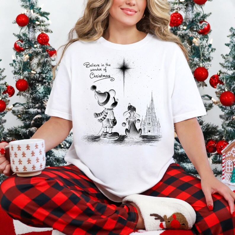 May include: White Christmas-themed t-shirt with a black and white illustration of cartoon characters and the text "Believe in the wonder of Christmas." The design includes a star, a castle, and two characters wearing winter attire.