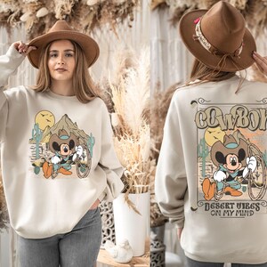May include: A beige sweatshirt with a cartoon image of Mickey Mouse dressed as a cowboy. The text "COWBOY" is printed in brown above the image and "DESERT VIBES ON MY MIND" is printed in brown below the image.