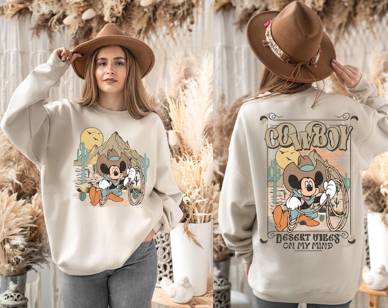 May include: A beige sweatshirt with a cartoon image of Mickey Mouse dressed as a cowboy. The text "COWBOY" is printed in brown above the image and "DESERT VIBES ON MY MIND" is printed in brown below the image.