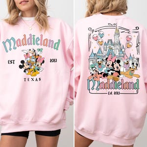 May include: Pink sweatshirt with a graphic of Mickey Mouse and friends in front of a castle. The text "Maddieland Est. 2023 Texas" is printed on the front of the sweatshirt. The back of the sweatshirt has a graphic of Mickey Mouse and friends in front of a castle with the text "Maddieland Est. 2023".
