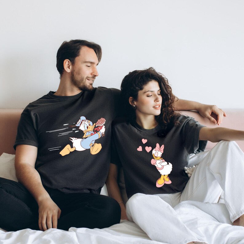 May include: A couple wearing matching black t-shirts with a cartoon duck design. The man's shirt features a duck holding flowers and running, while the woman's shirt features a duck blowing hearts.