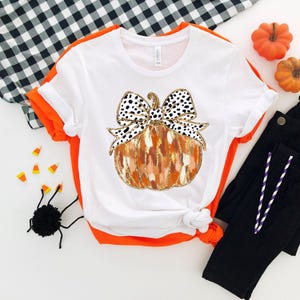 Puede incluir: Camiseta blanca con un gr&aacute;fico de una calabaza con un lazo de lunares negros y blancos. La calabaza es naranja y marr&oacute;n con detalles de purpurina dorada.