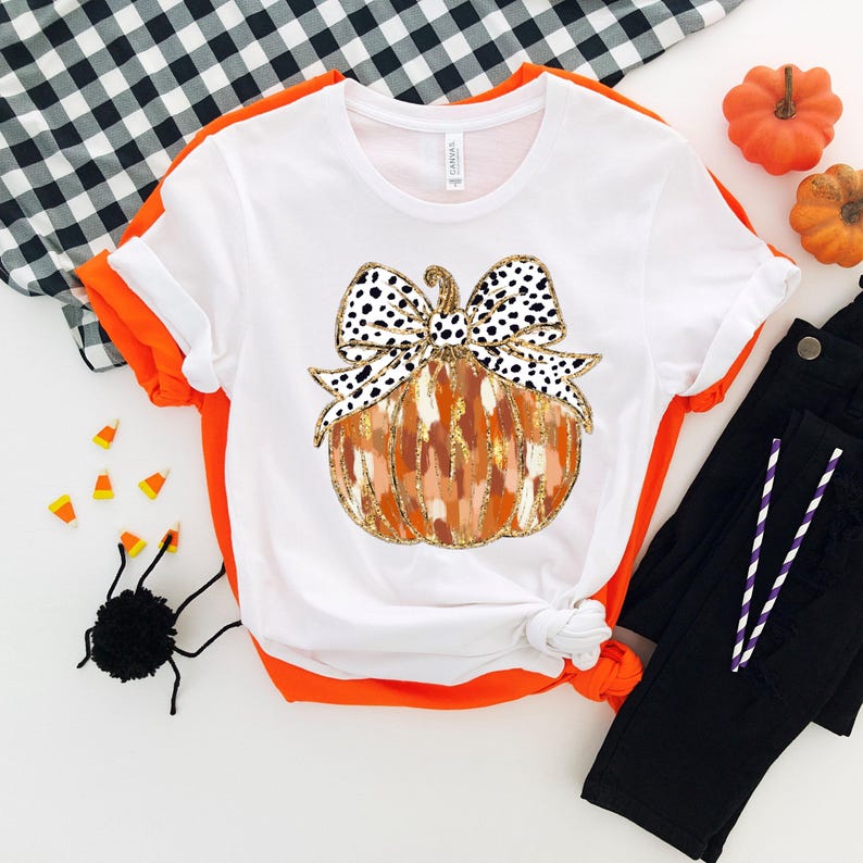 Puede incluir: Camiseta blanca con un gr&aacute;fico de una calabaza con un lazo de lunares negros y blancos. La calabaza es naranja y marr&oacute;n con detalles de purpurina dorada.