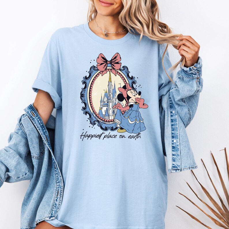 May include: Light blue t-shirt with a graphic of Minnie Mouse standing in front of Cinderella's Castle in a decorative frame. The text "Happiest place on earth" is printed below the graphic.