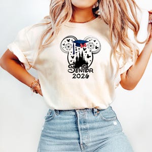 Puede incluir: Camiseta color crema con un dise&ntilde;o negro que presenta la forma de la cabeza de Mickey Mouse, un gorro de graduaci&oacute;n, fuegos artificiales y la silueta de un castillo. El texto "Senior 2026" est&aacute; impreso debajo del castillo.
