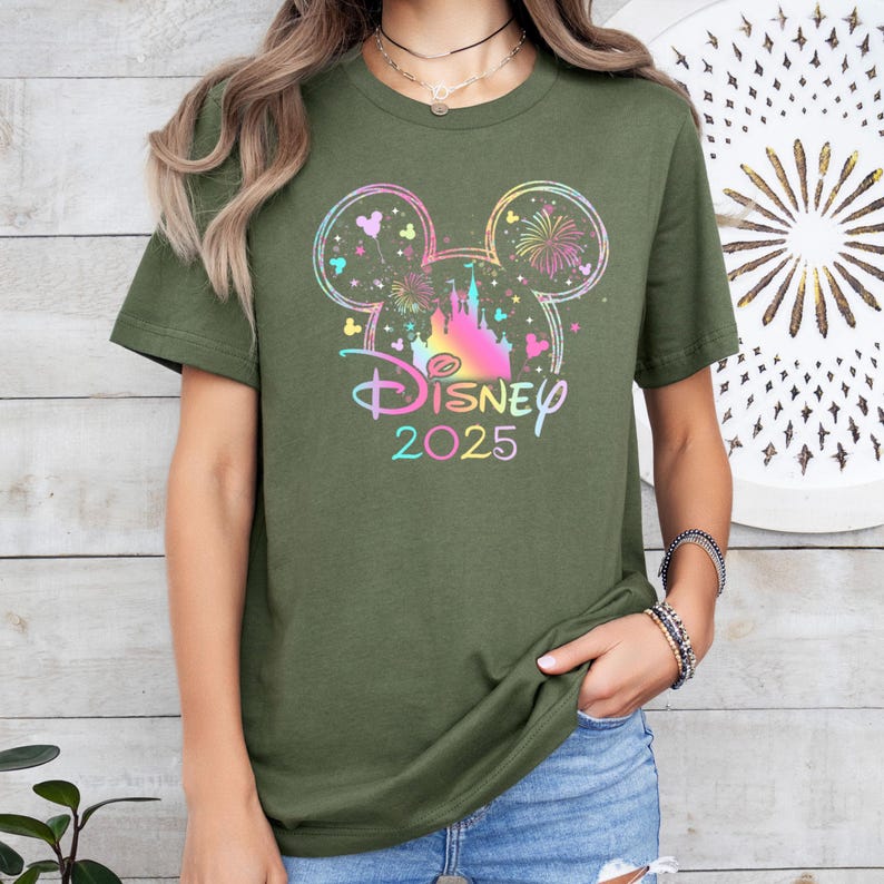 May include: A green t-shirt with a rainbow-colored Mickey Mouse head silhouette featuring a castle and fireworks. The text "DISNEY 2025" is printed below the silhouette.