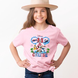 May include: A light pink t-shirt featuring a Disney Cruise design. The graphic includes Mickey and Minnie Mouse, a cruise ship, and the text "2025 Cruise". The design is in shades of red, blue, and white.