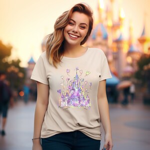 May include: A light beige t-shirt featuring a colourful illustration of a castle with balloons. The castle is outlined in black and filled with shades of purple, pink, blue, and green. The balloons are white with pink, yellow, and blue accents.