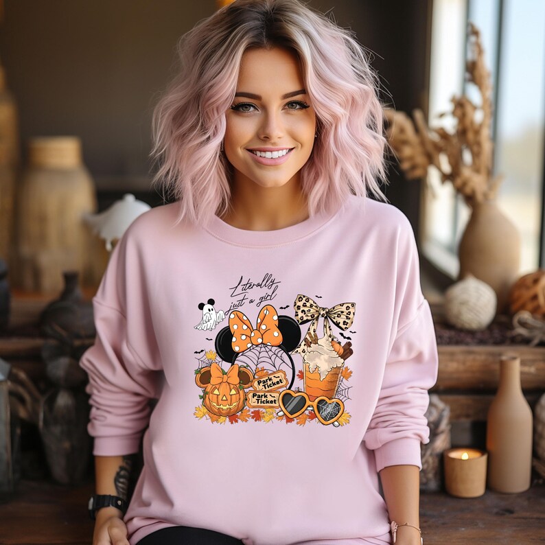May include: A light pink sweatshirt with a Halloween-themed graphic. The graphic includes a Minnie Mouse design with a pumpkin, spiderwebs, and the text "Literally just a girl". The design also features a ghost, sunglasses, and a beverage.