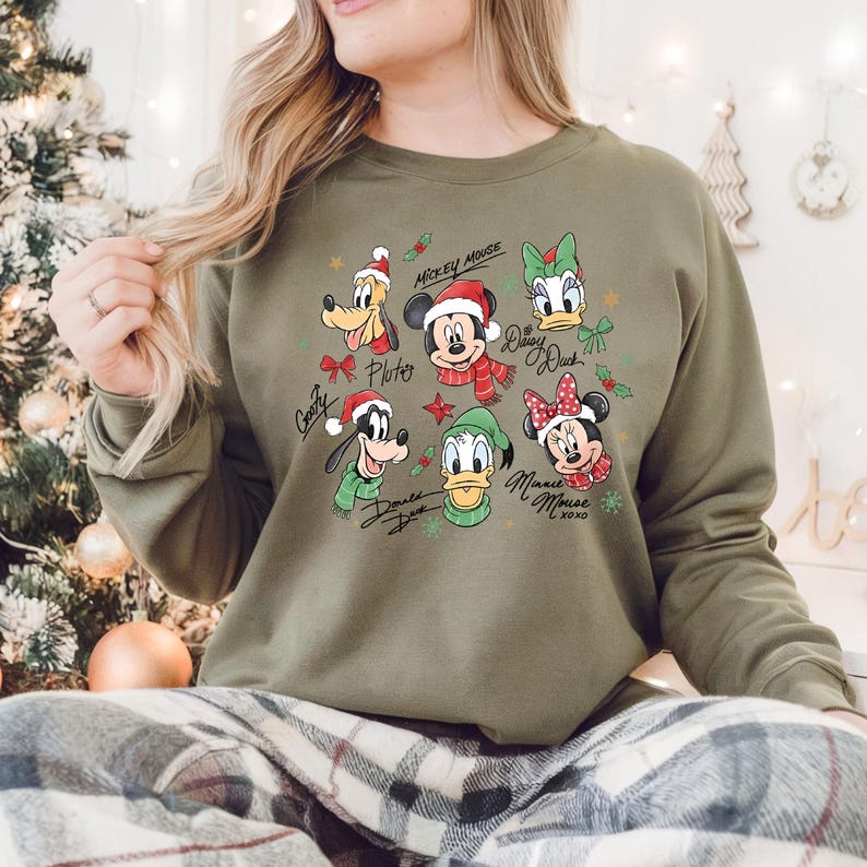 Może przedstawiać: Oliwkowo-zielona bluza z postaciami z kresk&oacute;wek Myszki Miki, Pluto, Goofy, Kaczora Donalda, Daisy Duck i Myszki Minnie, wszyscy w świątecznych strojach. Imiona postaci są napisane pod nimi czcionką skryptową. Widoczne są r&oacute;wnież ostrokrzew i płatki śniegu.