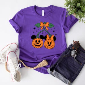 May include: A purple t-shirt featuring a Halloween-themed design. The graphic includes two smiling jack-o'-lanterns with mouse ears, connected by green vines and an orange bow. The shirt is paired with denim shorts and white sneakers.