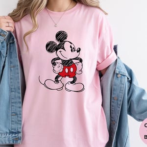 May include: A light pink t-shirt featuring a black and red sketch of Mickey Mouse. The cartoon character is standing with his hands on his hips. The shirt has rolled-up sleeves and is paired with a blue denim jacket.