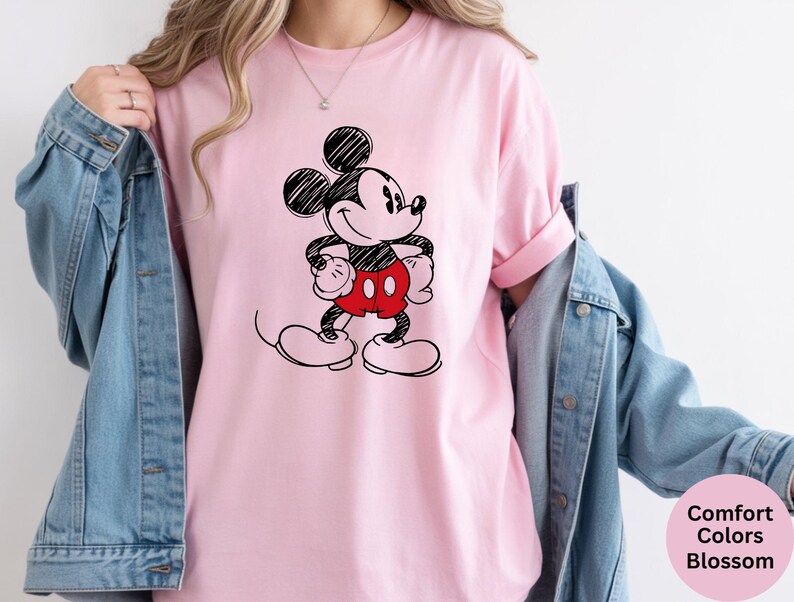 May include: A light pink t-shirt featuring a black and red sketch of Mickey Mouse. The cartoon character is standing with his hands on his hips. The shirt has rolled-up sleeves and is paired with a blue denim jacket.
