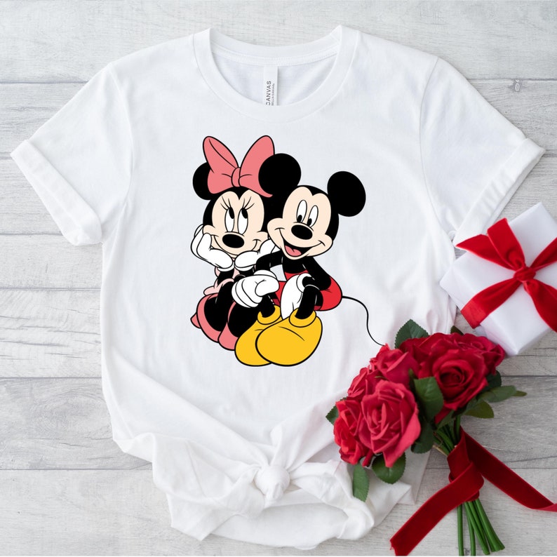 May include: White t-shirt with a graphic of Mickey Mouse and Minnie Mouse holding hands. The graphic is in black, red, and yellow.