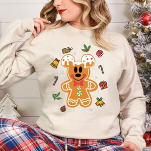 May include: A beige sweatshirt with a gingerbread Mickey Mouse design. The gingerbread Mickey Mouse is decorated with green and white icing and red candy. The sweatshirt has a festive Christmas theme with candy canes, bells, and holly.