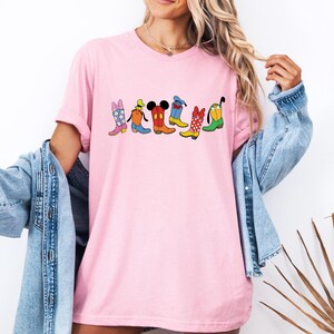 May include: Pink t-shirt featuring a colorful graphic of cartoon cowboy boots. The boots are in various colors and patterns, including floral, polka dots, and character-themed designs. The shirt has short sleeves and a relaxed fit.