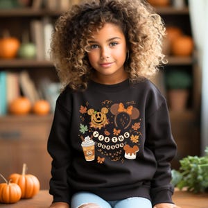 May include: A black sweatshirt featuring a fall-themed graphic with a pumpkin Mickey Mouse, Minnie Mouse ears, and a pumpkin spice latte. The text on the sweatshirt reads "'Tis the Season". The design includes fall leaves and a bear-shaped cookie.