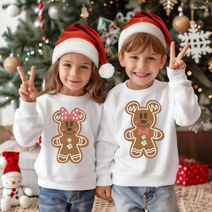 May include: Two children wearing white Christmas sweaters with gingerbread cookie designs. The girl's sweater features a gingerbread Minnie Mouse and the boy's sweater features a gingerbread Mickey Mouse. Both children are wearing red Santa hats.