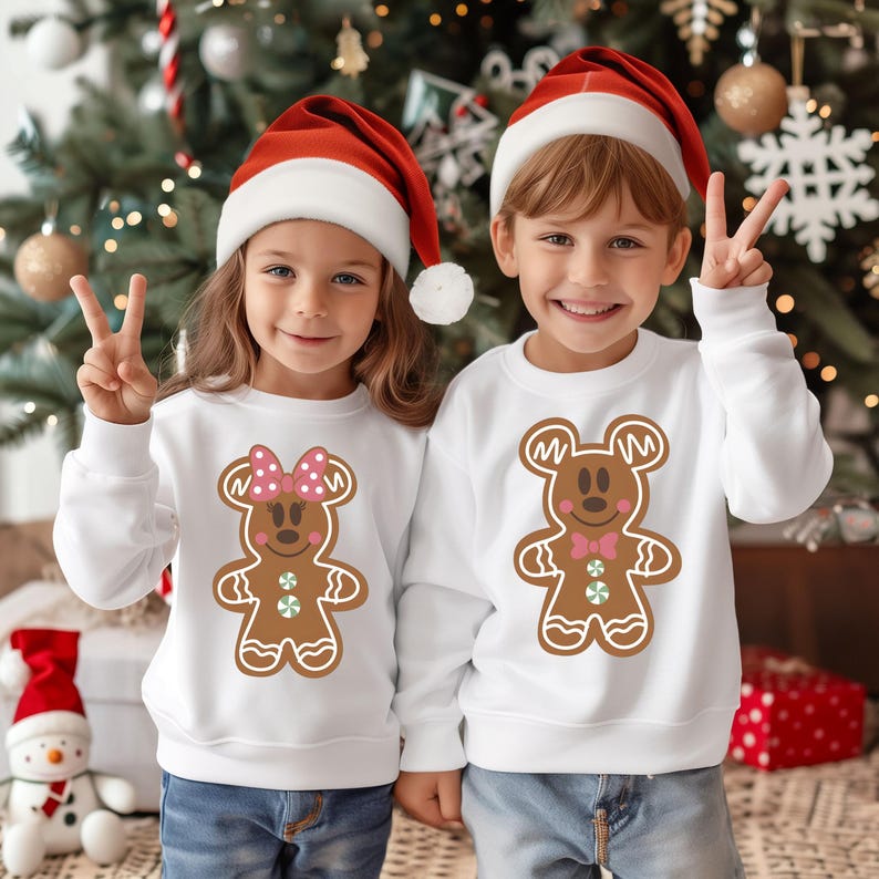 May include: Two children wearing white Christmas sweaters with gingerbread cookie designs. The girl's sweater features a gingerbread Minnie Mouse and the boy's sweater features a gingerbread Mickey Mouse. Both children are wearing red Santa hats.