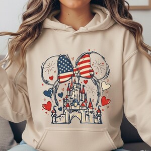 May include: A cream-colored hoodie featuring a graphic of a castle with Mickey Mouse ears, American flags, fireworks, and hearts in red, white, and blue. The design is a patriotic theme.