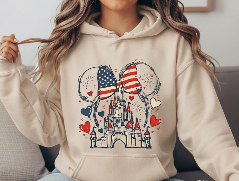 May include: A cream-colored hoodie featuring a graphic of a castle with Mickey Mouse ears, American flags, fireworks, and hearts in red, white, and blue. The design is a patriotic theme.