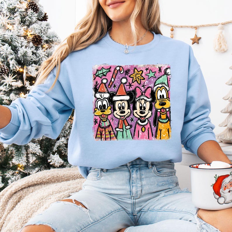 Op de afbeelding: Lichtblauw sweatshirt met een kleurrijke afbeelding van Disney-personages in kerstkleding. De afbeelding toont Mickey Mouse, Minnie Mouse, Donald Duck en Pluto, met feestelijke hoeden en decoraties. Het sweatshirt heeft lange mouwen.
